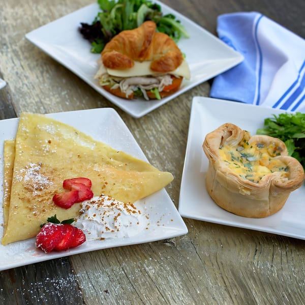 A sweet crepe with strawberries and whipped cream, a croissant sandwich, and a quiche on a wooden table.
