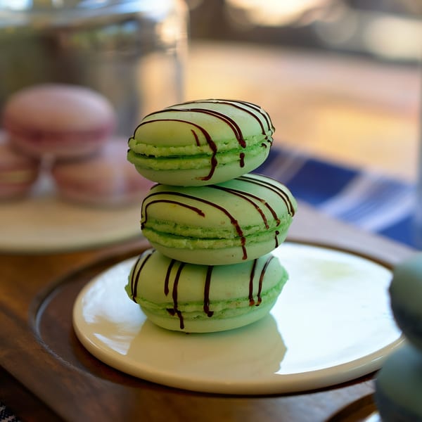 Stack of three green macarons drizzled with chocolate, on a white plate at The Crepe Club.