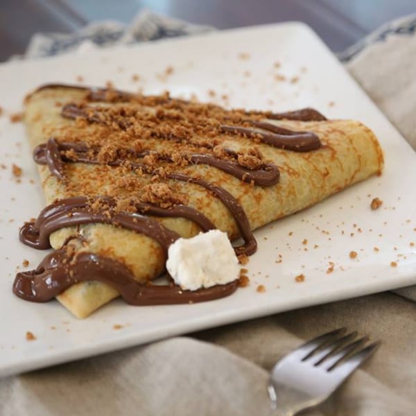 A golden crepe drizzled with chocolate and sprinkled with crumbs, served on a white plate.