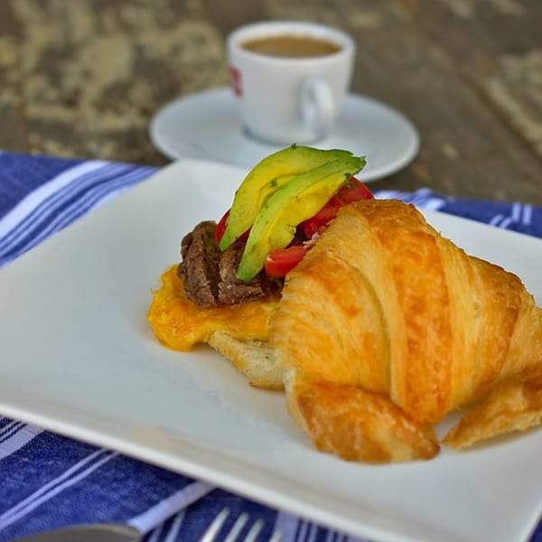 Savory croissant with meat, melted cheese, avocado, and tomatoes, with a coffee cup in the background.