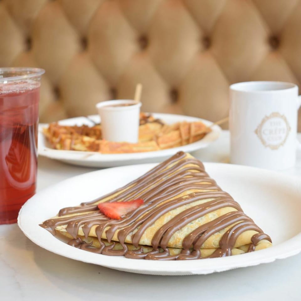A chocolate-drizzled crepe with a strawberry slice, drink, and coffee mug on a white table.