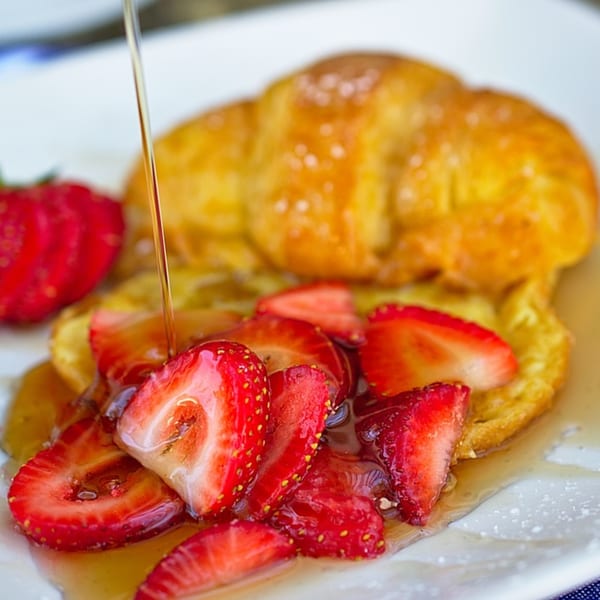 Maple syrup drizzling over sliced strawberries and a golden croissant on a white plate.