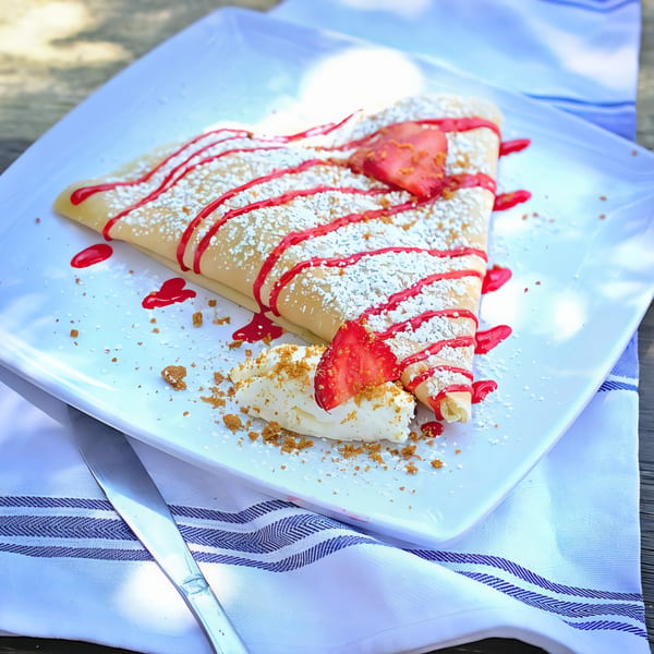 A strawberry crepe with powdered sugar, red drizzle, whipped cream, and graham cracker crumbles.