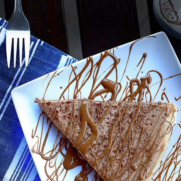 Chocolate crepe drizzled with chocolate sauce on a white plate with a fork and blue napkin.