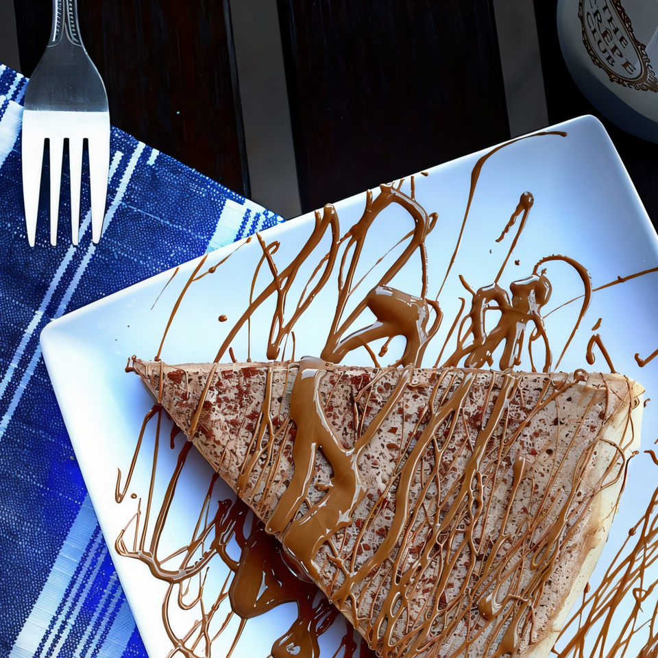Chocolate crepe drizzled with chocolate sauce on a white plate with a fork and blue napkin.