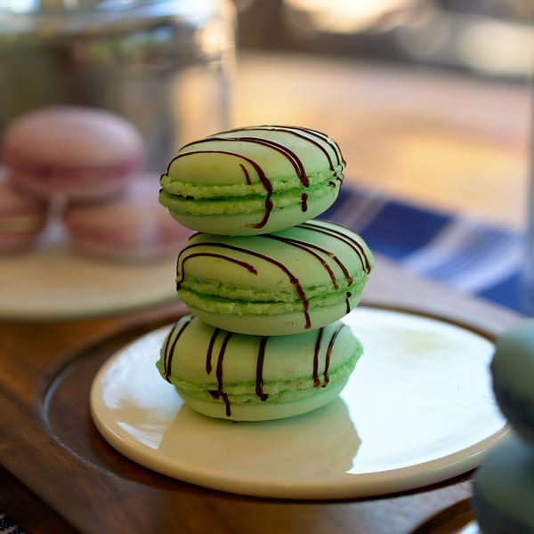 Three green macarons with chocolate drizzle stacked on a white plate at The Crepe Club.