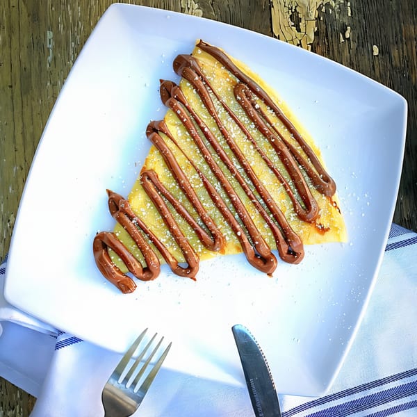 A golden crepe drizzled with chocolate sauce and powdered sugar, served on a white plate.