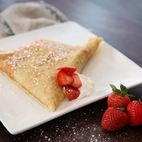 A golden crepe dusted with powdered sugar, served with whipped cream and fresh strawberries on a white plate.