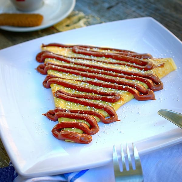 A golden crepe drizzled with chocolate sauce and powdered sugar on a white plate.