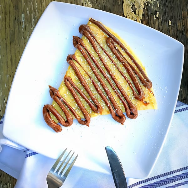 A golden crepe drizzled with chocolate and powdered sugar on a white plate with cutlery.