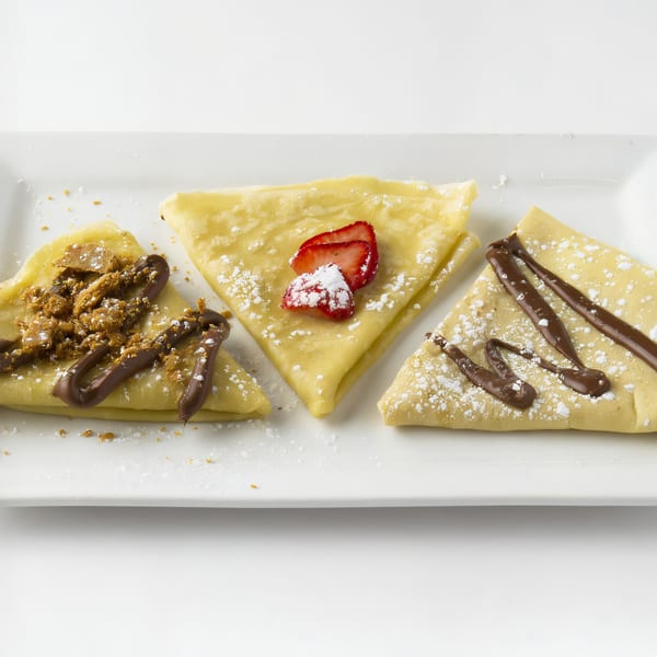 Three crepes on a white plate, one with strawberries, one with chocolate and crumbs, and one with chocolate drizzle.