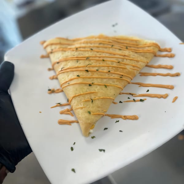 A hand in a black glove holds a white plate with a folded crepe drizzled with orange sauce and herbs.
