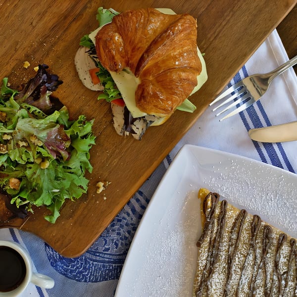 Savory croissant sandwich with salad, sweet crepe, and coffee on a wooden board and blue napkin.